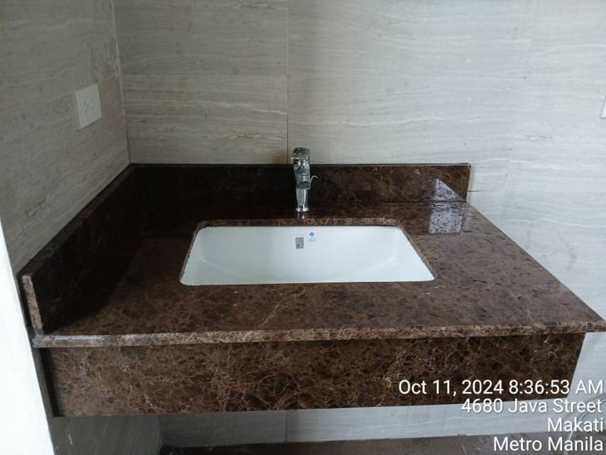 A brown marble bathroom countertop with a white rectangular sink and a chrome faucet, set against light-colored tiled walls. Date, time, and location text is visible in the bottom right corner.