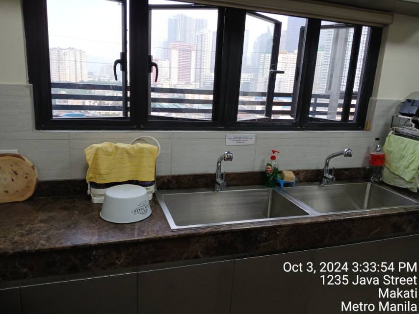 A kitchen counter with two sinks, a cutting board, a drying rack, and cleaning supplies; buildings and cityscape visible through the window. Date and location text is overlaid.