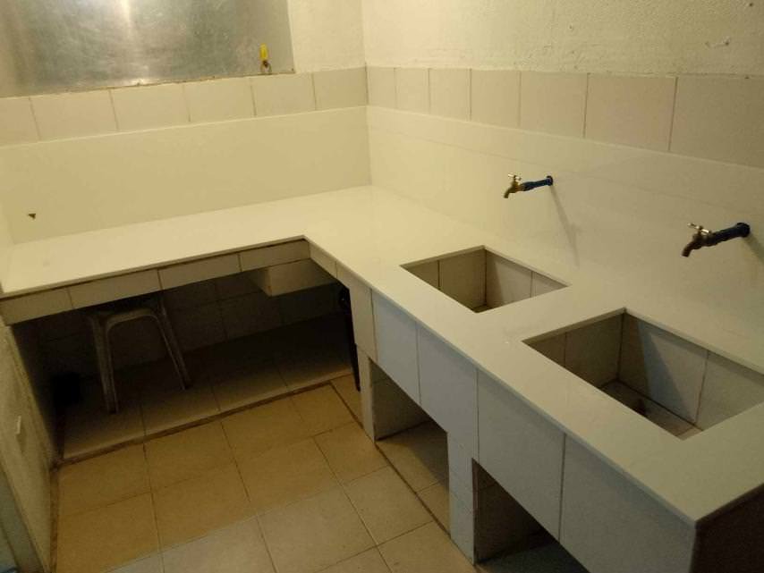 A tiled kitchen countertop with two sink cutouts and two wall-mounted faucets, next to a corner workspace with a metal stool underneath.